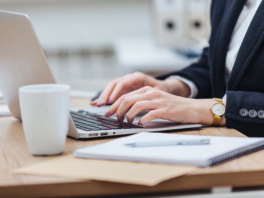 Closeup of woman's hands typing on a laptop in an office with a mug, notepad, and pen on the desk