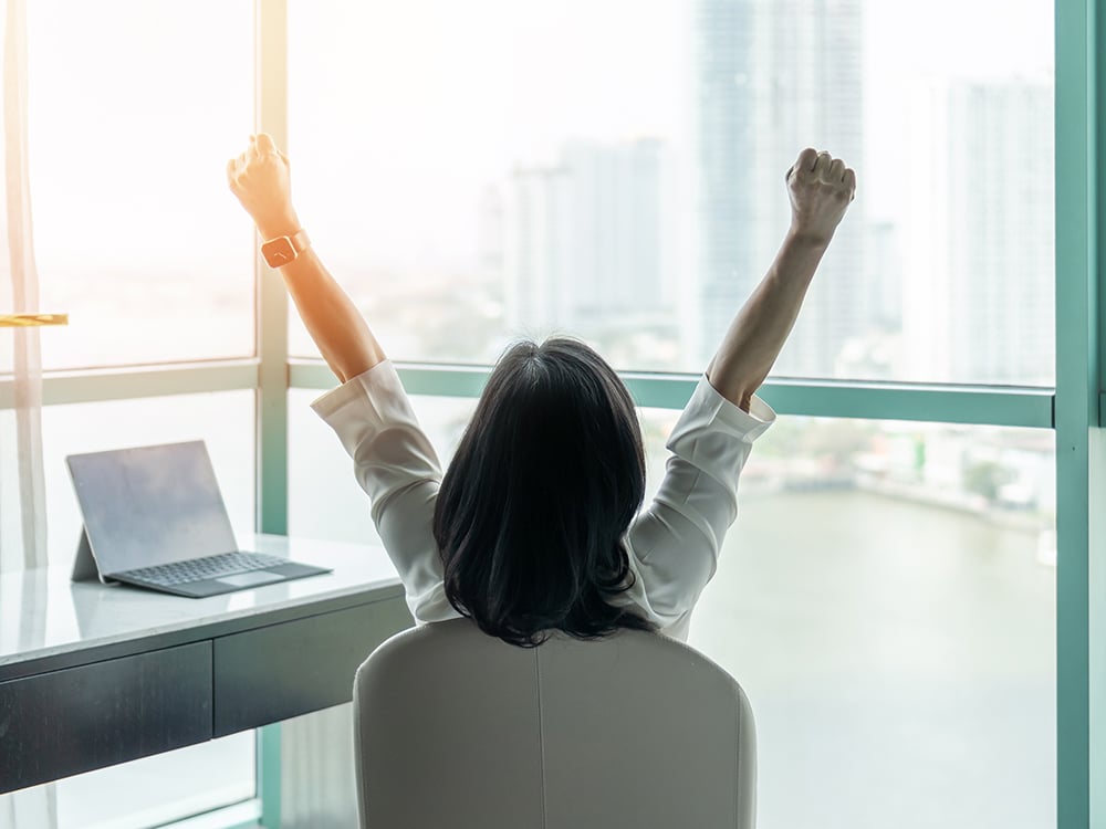 Happy businesswoman with her arms up sitting at a desk and looking out the window at a city skyline