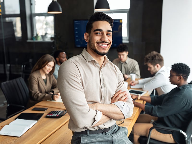 Young bearded businessman sitting on desk and posing as coworkers collaborate in background