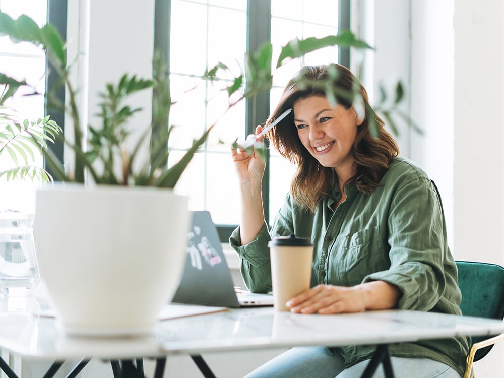 Young smiling woman working on a laptop, holding a cup of coffee, with a house plant in the foreground