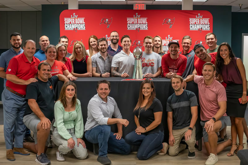 Group of employees gathered around the Lombardi Trophy in front of a Tampa Bay Buccaneers Super Bowl champions backdrop